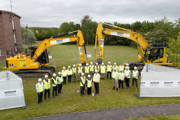 A group of people in high-visibility vests and helmets, including Royal Engineers, stand in front of two large diggers on a grassy area at Catterick, with Morgan Sindall construction fencing and signage visible.