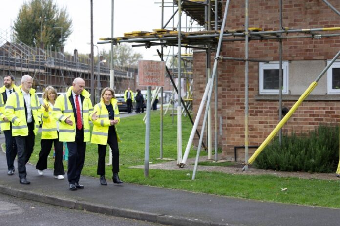 A group of people in high-visibility jackets walk past scaffolding and a brick building on a residential street, inspecting military homes as part of the Defence Housing Service initiative.