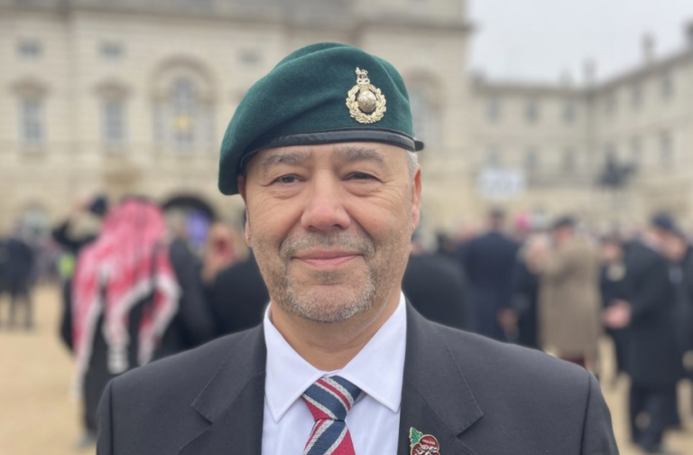 A man wearing a green beret, suit, and striped tie stands outdoors with people and a historic building in the background, possibly attending a Blind Veterans UK event in Holderness.