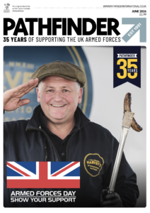 A smiling man in a cap and black jacket holds a grilled sausage with tongs. UK flag and