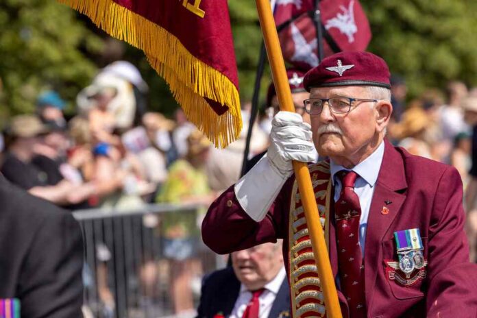 An elderly man in a maroon blazer and beret holds a flag during an outdoor event, wearing medals and gloves, with a crowd and metal barrier in the background.