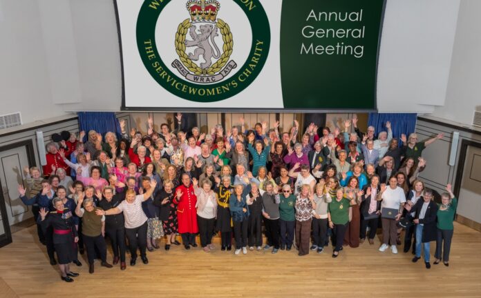 A large group of people pose and wave for a photo at the WRAC Association Annual General Meeting in a hall with a charity logo and event sign behind them.