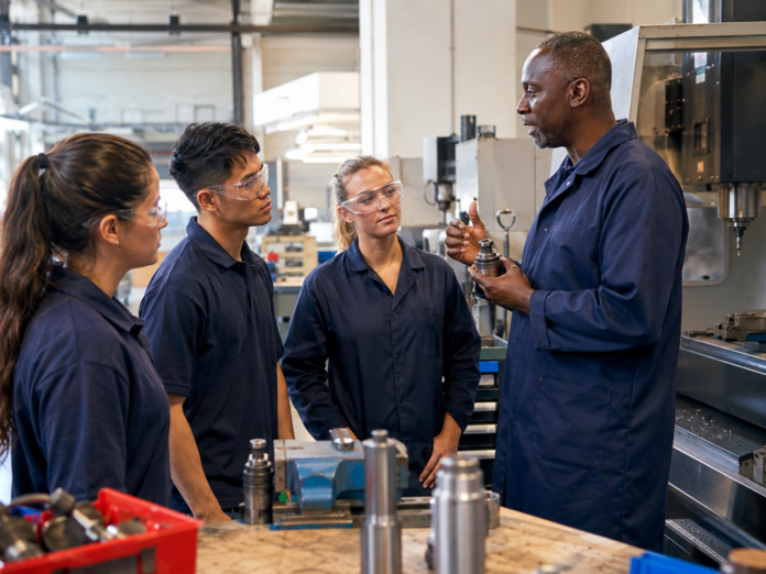 A man provides training on a metal tool to three colleagues in safety glasses and work uniforms inside a factory or workshop with machinery.