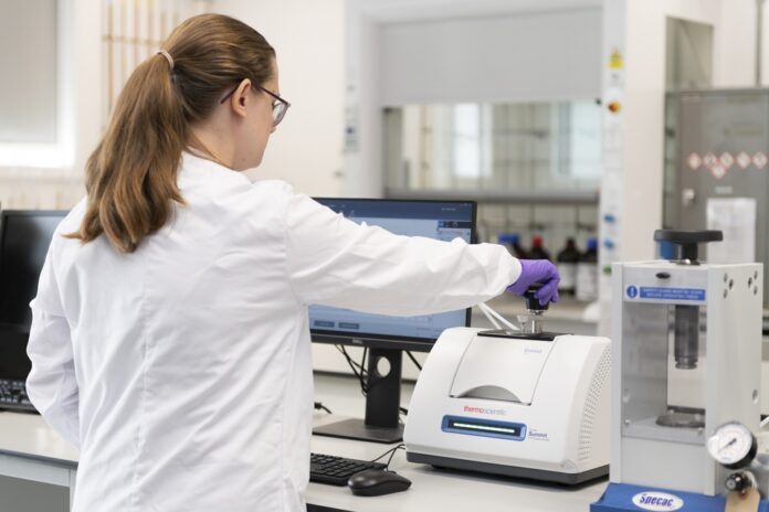 A person in a lab coat and gloves operates a lab machine in a laboratory, with a computer monitor and various equipment visible on the bench.